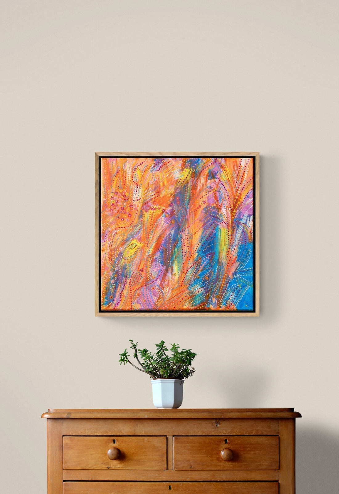 Framed abstract painting with vibrant orange, blue, and pink brushstrokes hanging above a wooden cabinet with a potted plant